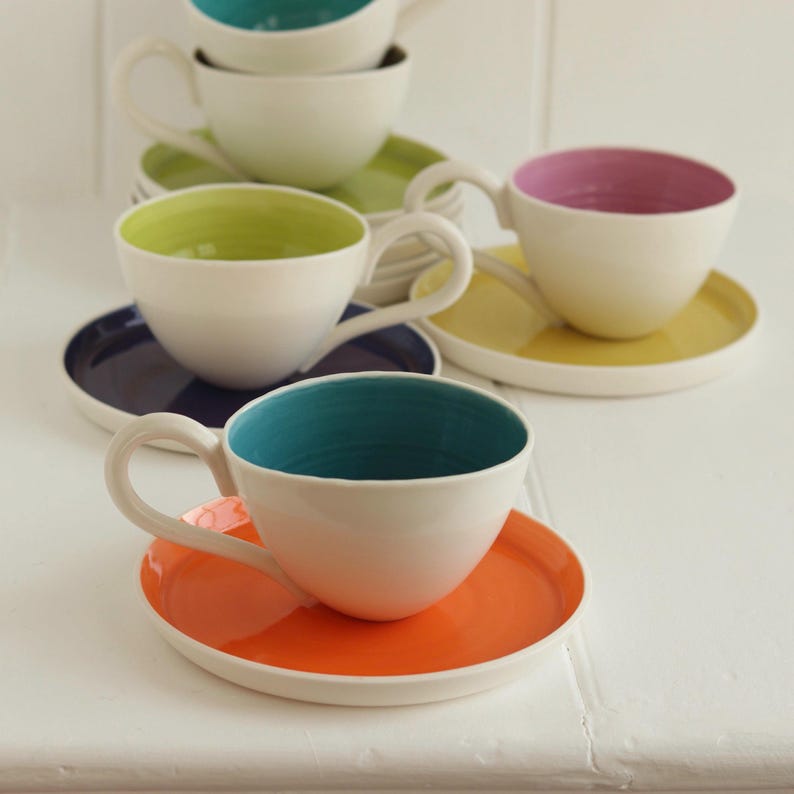 May include: A collection of white ceramic teacups and saucers with brightly colored interiors and matching saucers. The teacups have teal, green, pink, and orange interiors, with corresponding saucers in the same colors. The teacups have curved handles.