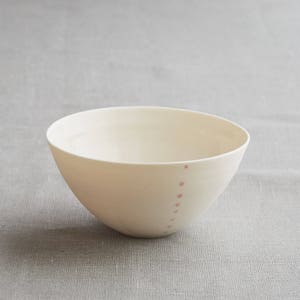 Może przedstawiać: Kremowa ceramiczna miska o prostym, nowoczesnym designie. Miska ma zaokrąglony kształt i szereg małych, r&oacute;żowych kropek biegnących wzdłuż boku. Miska jest ustawiona na neutralnym szarym tle.
