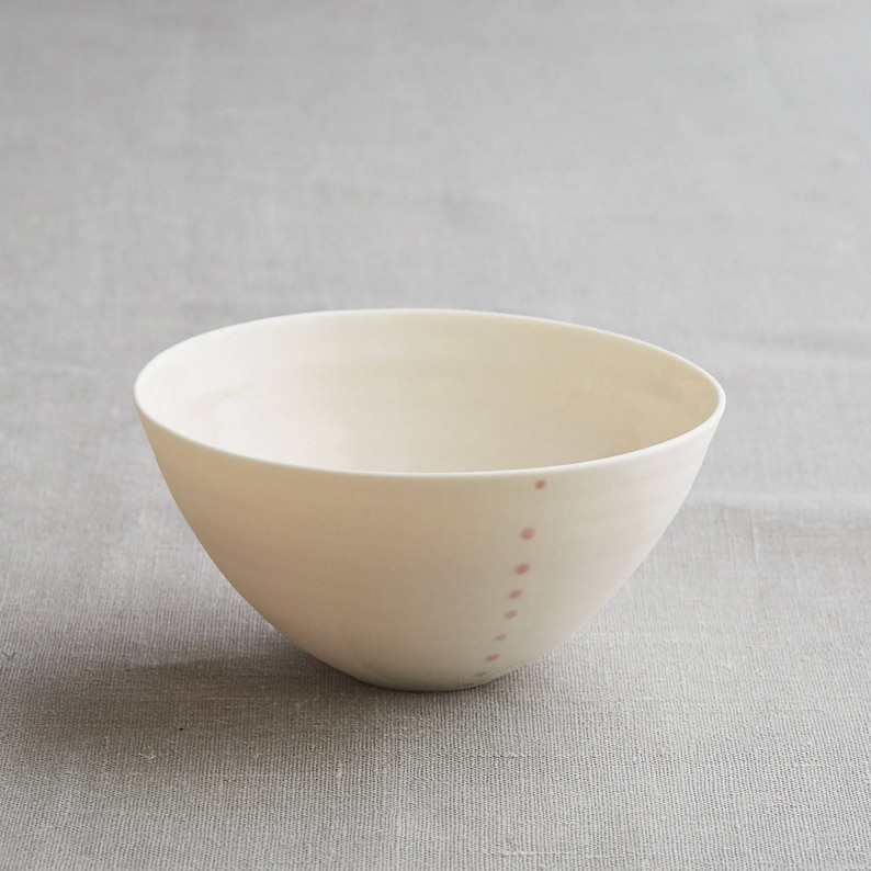 Może przedstawiać: Kremowa ceramiczna miska o prostym, nowoczesnym designie. Miska ma zaokrąglony kształt i szereg małych, r&oacute;żowych kropek biegnących wzdłuż boku. Miska jest ustawiona na neutralnym szarym tle.