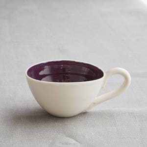 May include: A white ceramic teacup with a deep purple interior and a curved handle. The cup is set against a gray background, highlighting its simple, elegant design. The teacup is ideal for serving tea or coffee.