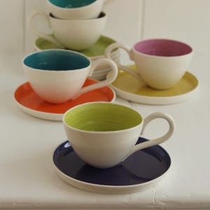 May include: A collection of white teacups with colorful interiors and matching saucers. The teacups have teal, pink, green, and yellow interiors, and the saucers are orange, yellow, green, and navy blue. The cups have curved handles.