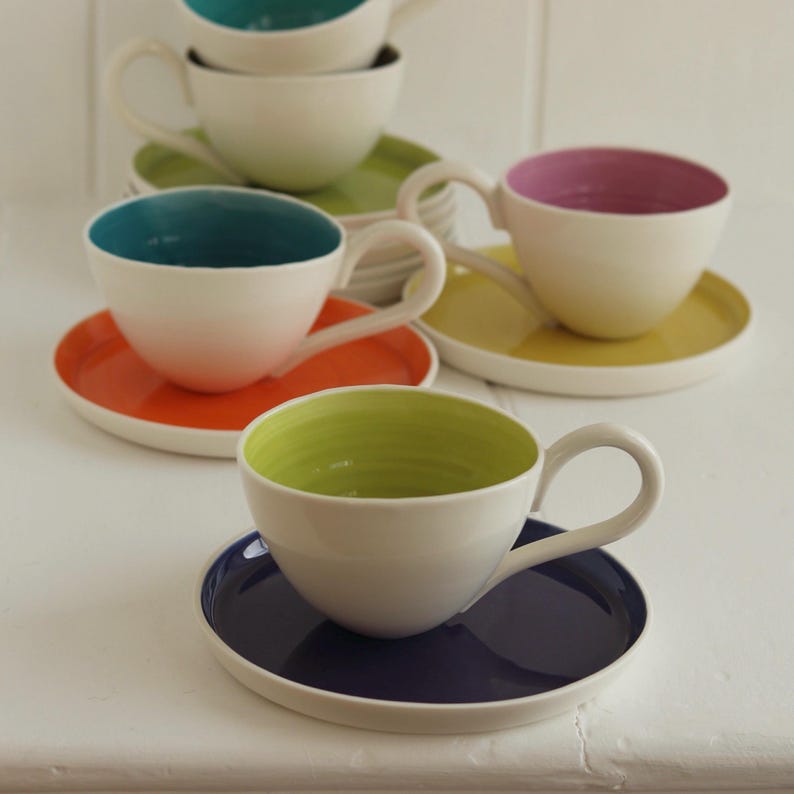 May include: A collection of white teacups with colorful interiors and matching saucers. The teacups have teal, pink, green, and yellow interiors, and the saucers are orange, yellow, green, and navy blue. The cups have curved handles.