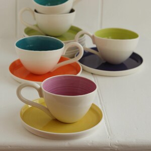 May include: A collection of white ceramic teacups and saucers with colorful interiors and matching saucers. The teacups have interiors in teal, lime green, orange, and pink. The saucers are in coordinating colors, creating a vibrant and modern aesthetic.