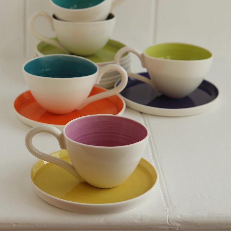 May include: A collection of white ceramic teacups and saucers with colorful interiors and matching saucers. The teacups have interiors in teal, lime green, orange, and pink. The saucers are in coordinating colors, creating a vibrant and modern aesthetic.