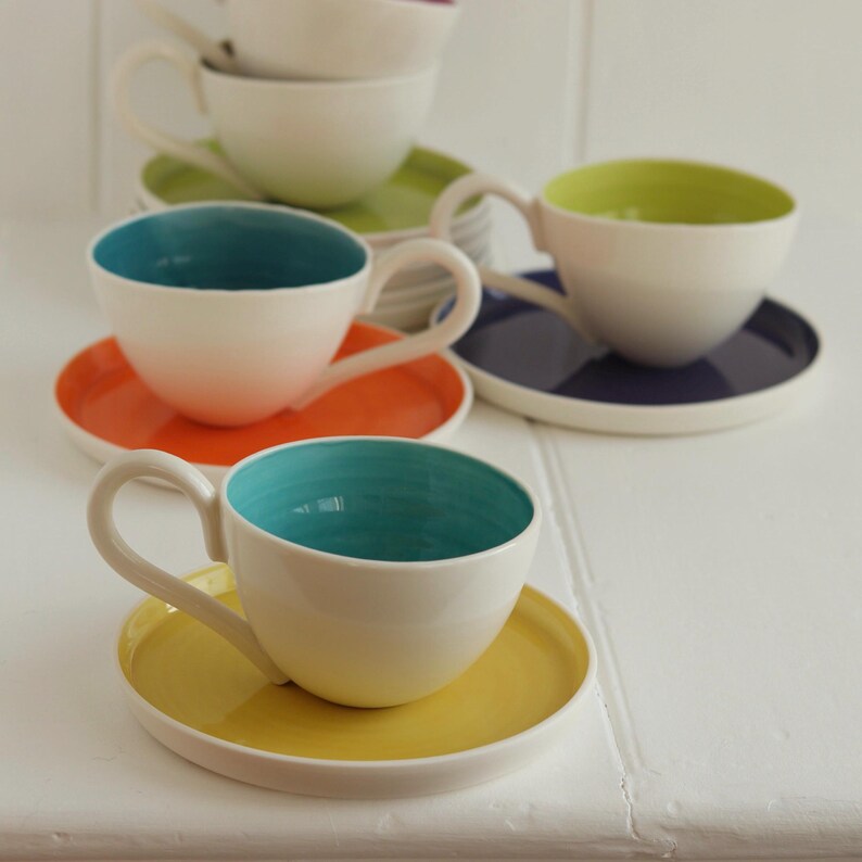May include: A collection of white ceramic teacups with colorful interiors and matching saucers. The teacups have turquoise, lime green, orange, and yellow interiors, with corresponding saucers. The teacups are arranged on a white surface.