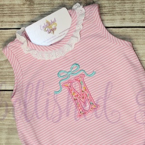 May include: Pink and white striped baby romper with a floral embroidered letter "M" and a light blue bow. The romper has a white lace trim around the neckline and a tag attached at the top.