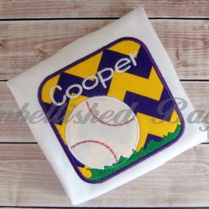 May include: White t-shirt with an embroidered patch featuring a baseball and the name "Cooper" in a yellow and purple chevron pattern.