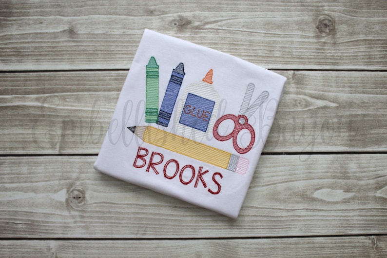 K&ouml;nnte beinhalten: Wei&szlig;es T-Shirt mit einem roten, gelben, gr&uuml;nen und blauen Buntstift, einer roten Schere, einer wei&szlig;en Klebeflasche und einem gelben Bleistift mit dem in Rot gestickten Wort "BROOKS".