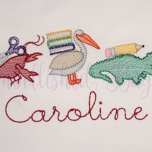 Puede incluir: Dise&ntilde;o bordado con un langostino rojo sosteniendo tijeras, un pel&iacute;cano cargando una pila de libros, un cocodrilo verde con un l&aacute;piz amarillo en la espalda y el nombre "Caroline" en hilo rojo.