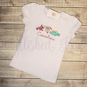 Puede incluir: Camiseta blanca de manga corta con un dise&ntilde;o bordado de un pel&iacute;cano, un cangrejo, un l&aacute;piz y un caim&aacute;n. El nombre "Caroline" est&aacute; bordado debajo del dise&ntilde;o.