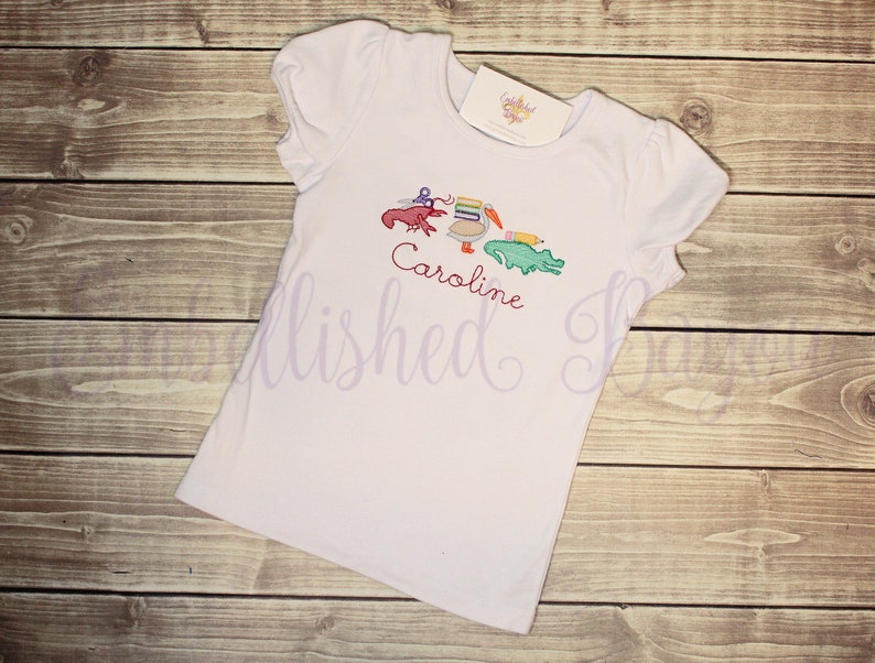 Puede incluir: Camiseta blanca de manga corta con un dise&ntilde;o bordado de un pel&iacute;cano, un cangrejo, un l&aacute;piz y un caim&aacute;n. El nombre "Caroline" est&aacute; bordado debajo del dise&ntilde;o.