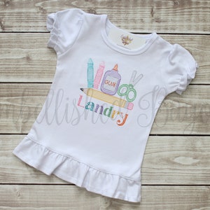 K&ouml;nnte beinhalten: Wei&szlig;es T-Shirt mit kurzen &Auml;rmeln und einem R&uuml;schen-Saum. Das Shirt hat ein gesticktes Design von Schulutensilien, darunter ein Bleistift, Buntstifte, eine Schere und Kleber, mit dem Namen "Landry" darunter gestickt.