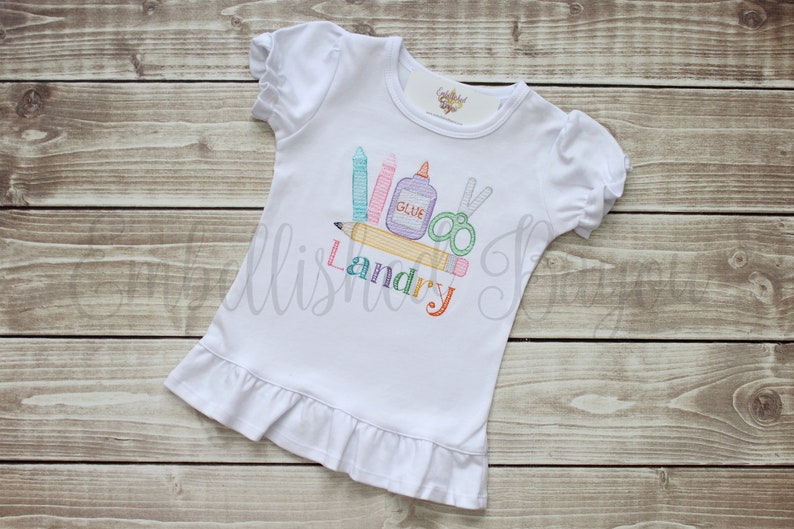 K&ouml;nnte beinhalten: Wei&szlig;es T-Shirt mit kurzen &Auml;rmeln und einem R&uuml;schen-Saum. Das Shirt hat ein gesticktes Design von Schulutensilien, darunter ein Bleistift, Buntstifte, eine Schere und Kleber, mit dem Namen "Landry" darunter gestickt.