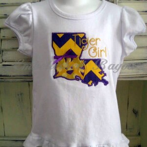 May include: White t-shirt with a purple and yellow embroidered design of the state of Louisiana with the words "Tiger Girl".