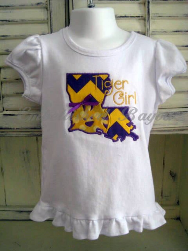 May include: White t-shirt with a purple and yellow embroidered design of the state of Louisiana with the words "Tiger Girl".