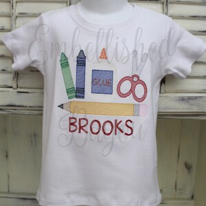 K&ouml;nnte beinhalten: Wei&szlig;es T-Shirt mit einem gestickten Design von Schulutensilien, darunter Buntstifte, Schere, Kleber und ein Bleistift. Der Text "BROOKS" ist unter den Utensilien gestickt.