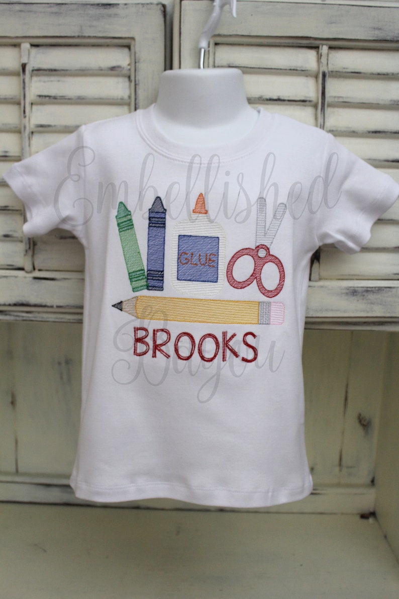 K&ouml;nnte beinhalten: Wei&szlig;es T-Shirt mit einem gestickten Design von Schulutensilien, darunter Buntstifte, Schere, Kleber und ein Bleistift. Der Text "BROOKS" ist unter den Utensilien gestickt.