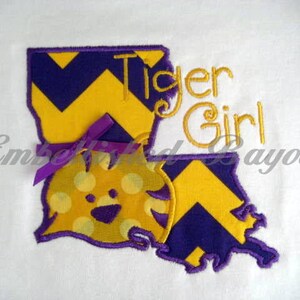 May include: An embroidered design of the state of Louisiana with a yellow and purple chevron pattern. The design includes the words "Tiger Girl" and a cartoon tiger with yellow and purple polka dots.