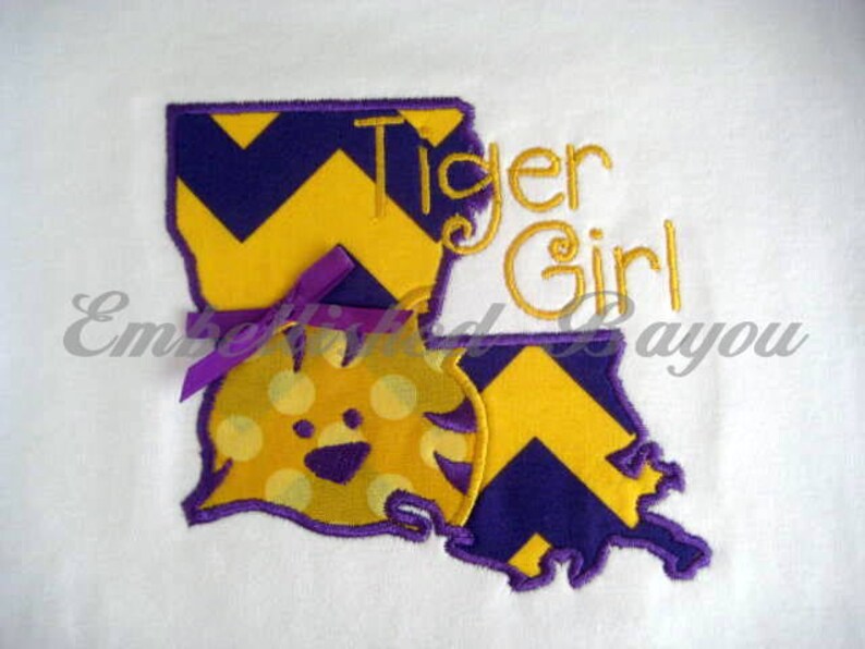 May include: An embroidered design of the state of Louisiana with a yellow and purple chevron pattern. The design includes the words "Tiger Girl" and a cartoon tiger with yellow and purple polka dots.
