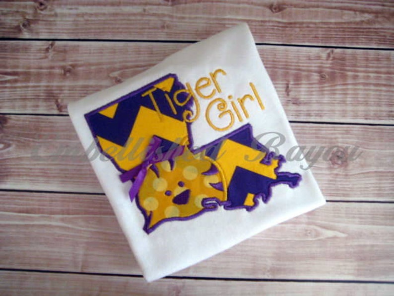 May include: White t-shirt with an embroidered design of a purple and gold chevron pattern, a yellow and white polka dot design, and the text "Tiger Girl".