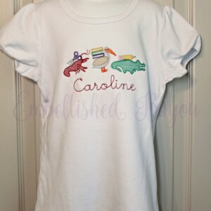 Puede incluir: Camiseta blanca de manga corta con un dise&ntilde;o bordado de un pel&iacute;cano que lleva libros, un cangrejo de r&iacute;o, un l&aacute;piz y un caim&aacute;n. El nombre "Caroline" est&aacute; bordado debajo del dise&ntilde;o.