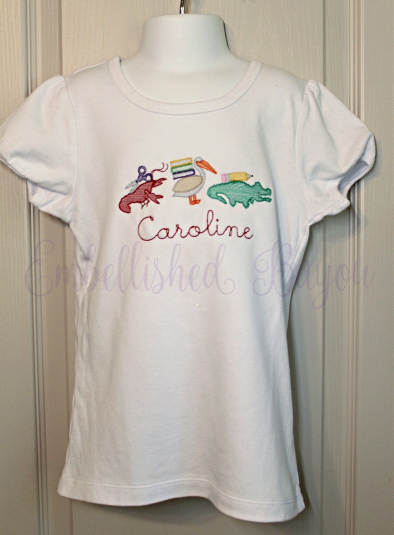 Puede incluir: Camiseta blanca de manga corta con un dise&ntilde;o bordado de un pel&iacute;cano que lleva libros, un cangrejo de r&iacute;o, un l&aacute;piz y un caim&aacute;n. El nombre "Caroline" est&aacute; bordado debajo del dise&ntilde;o.