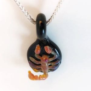 May include: A black glass pendant with a detailed orange and purple scorpion design. The pendant is attached to a silver chain.