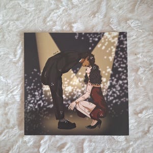 May include: Square art print featuring a couple in an embrace under stage lights. The man is in a black suit, and the woman is in a red dress. The background has a golden glow with white sparkles.
