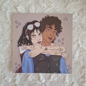 May include: Square art print featuring two figures embracing. One figure has dark hair and is wearing a blue vest. The other figure has dark hair and is wearing sunglasses. The background is a light purple color with star shapes.