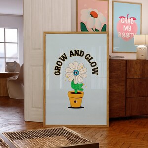 Peut inclure: Une affiche bleu clair avec une fleur blanche dans un pot jaune. La fleur a un œil bleu et des paillettes dorées. Le texte "GROW AND GLOW" est écrit en arc noir et or autour de la fleur.