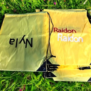 Op de afbeelding: Een stapel gele trekkoordtassen met zwarte trekkoorden. De bovenste tas heeft het woord "Raidon" in wit en rood gedrukt. De onderste tas heeft het woord "Nyle" in zwart gedrukt.