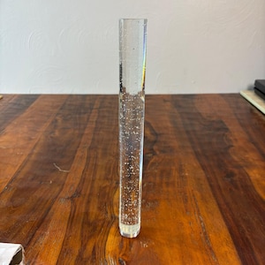 May include: A clear, rectangular acrylic rod with embedded bubbles. The rod is standing upright on a wooden surface. The bubbles are scattered throughout the clear material, creating a decorative effect. The rod is a decorative item.