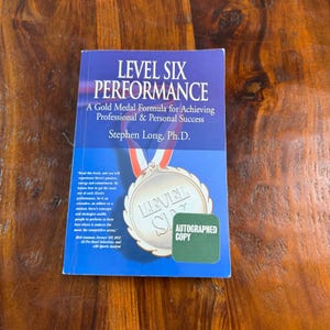 May include: A paperback book titled Level Six Performance: A Gold Medal Formula for Achieving Professional & Personal Success by Stephen Long, Ph.D. The blue cover features a graphic of a gold medal with a red, white, and blue ribbon and a green sticker.