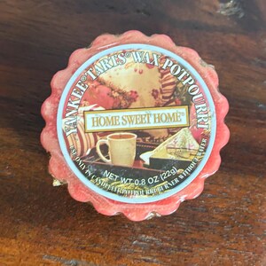 以下が含まれることがあります： 丸い赤いヤンキーキャンドルのワックスタルト、スカラップエッジ付き。「Yankee Tarts Wax Potpourri」と「Home Sweet Home」と書かれたラベル。正味重量は22gです。ラベルの画像にはマグカップとケーキのスライスが描かれています。
