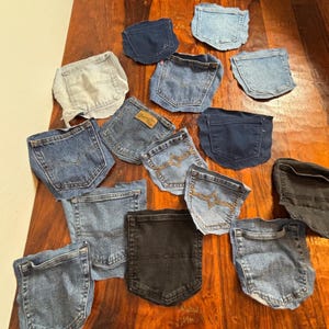 May include: A collection of denim pockets in various shades of blue and black, arranged on a wooden surface. The pockets are from jeans and have different stitching details and brand labels.