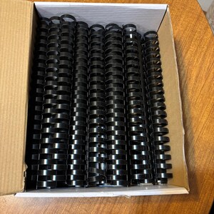 May include: A box containing numerous black plastic binding combs, ideal for document organisation. The combs are neatly stacked, ready for use. The dark colour of the combs contrasts with the light brown of the cardboard box.