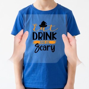 Puede incluir: Una camiseta azul real de manga corta con un icono de sombrero de bruja negro y el texto naranja y negro "Eat Drink and Be Scary" impreso en el pecho, centrado en una superposición gráfica rectangular transparente.
