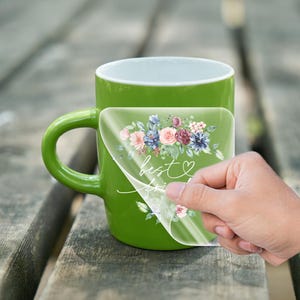 May include: A green mug with a clear sticker on the front. The sticker features a watercolor floral design with pink, blue, and purple flowers. The text "best friend" is written in white cursive on the sticker.