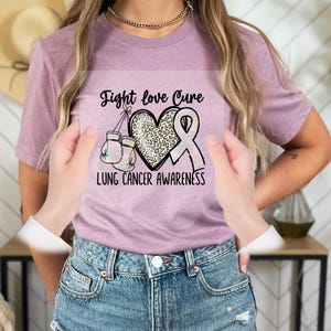 A person wearing a pink t-shirt with a graphic design related to lung cancer awareness, including a heart, boxing gloves, and a pink ribbon.