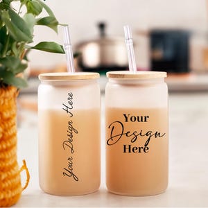 May include: Two frosted glass tumblers with bamboo lids and clear straws. The tumblers are filled with a light brown liquid and have the text "Your Design Here" printed on them.