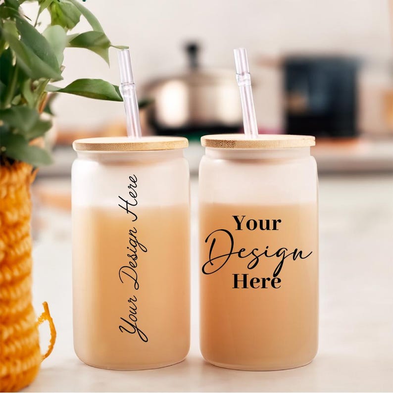May include: Two frosted glass tumblers with bamboo lids and clear straws. The tumblers are filled with a light brown liquid and have the text "Your Design Here" printed on them.