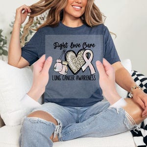 A woman wearing a blue t-shirt with a pink ribbon and the text "Fight love Cure lung cancer awareness" is sitting on a couch.