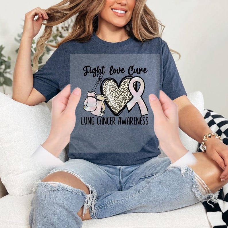A woman wearing a blue t-shirt with a pink ribbon and the text "Fight love Cure lung cancer awareness" is sitting on a couch.