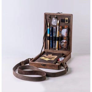 May include: A wooden box with a leather strap, containing various writing utensils, glasses, and a notebook. The box is open and the contents are visible.