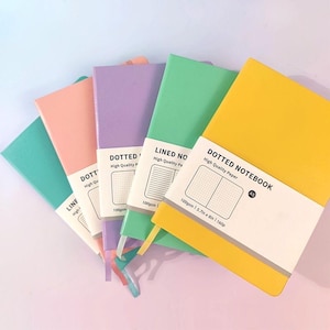 May include: Five A5 notebooks in pastel colours: teal, peach, lilac, green, and yellow. Each has a white band with the text "DOTTED NOTEBOOK" or "LINED NOTEBOOK". The notebooks are approximately 14.5cm x 20.3cm.