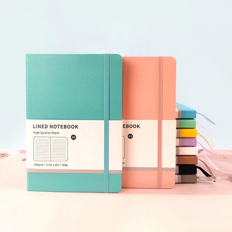 May include: Two A5 lined notebooks, one teal and one coral, with elastic closures. The teal notebook has the text "LINED NOTEBOOK" and "High Quality Paper". Each notebook is 5.7in x 8in and 160p.