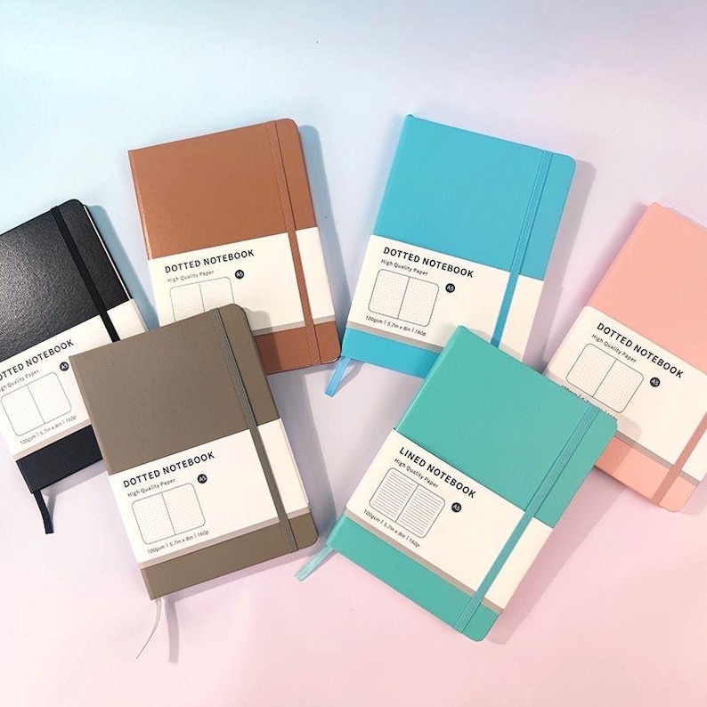 May include: A collection of colorful notebooks in various shades, including black, brown, teal, blue, pink, and gray. Each notebook has a white band with the text "DOTTED NOTEBOOK" or "LINED NOTEBOOK" and a diagram of the interior pages. The notebooks are A5 size.