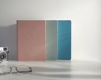 May include: Three hardcover journals in pastel colors: dusty rose, pale green, and teal. A vintage camera and eyeglasses are in the foreground. The journals are standing upright on a white surface against a white wall.