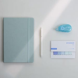 Può includere: Un quaderno azzurro con elastico, una penna bianca, un applicatore di punti blu e un pacco di post-it con linguette blu e viola. La parola "STICKER" è stampata sui post-it.