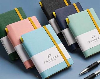 May include: A set of six notebooks in various colors, including green, pink, blue, and black. Each notebook has a yellow elastic band closure and a white label with the text "A7 便携式口袋笔记本 NOTEBOOK" in black. The notebooks are arranged in a staggered pattern on a light blue surface.